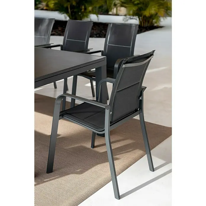 Sunfun Anna Sillón de jardín Anna - 56,5 x 67 x 91,5 cm, Antracita, Acero/Textiline - Silla de comedor de exterior gris oscuro con tela de malla negra, junto a una mesa a juego, sobre una alfombra de patio.