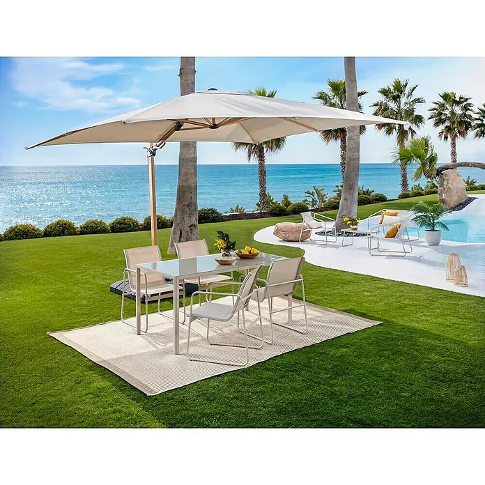 Sunfun Parasol de jardín Capri - Crudo, 300 x 350 cm, Aluminio - Conjunto de comedor exterior con mesa de cristal y cuatro sillas beige claro, sobre una alfombra bajo una gran sombrilla en jardín junto al mar.