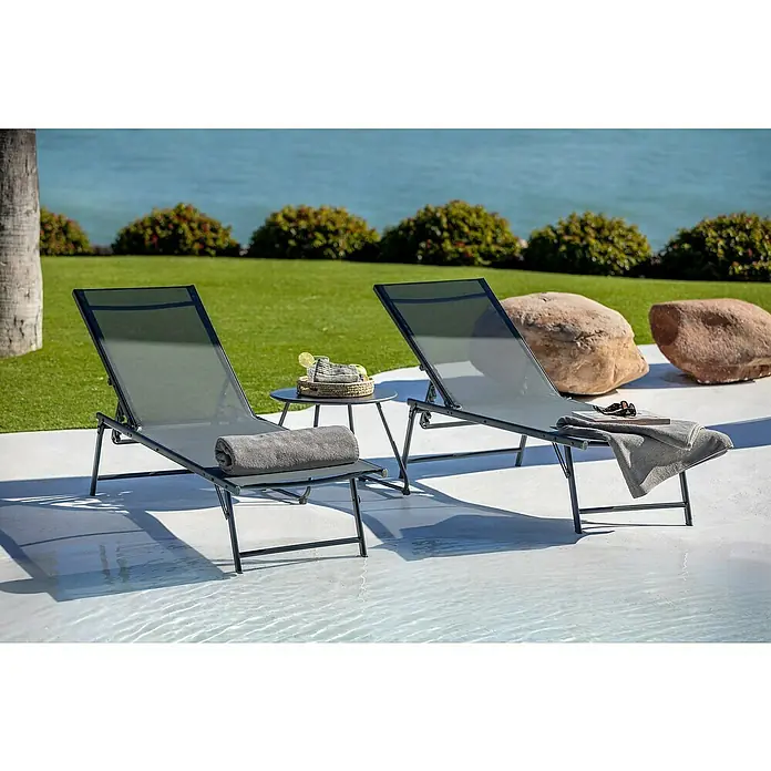 Tumbona de jardín Belisa - Gris oscuro, Acero, Abatible - Dos tumbonas gris oscuro y una mesa redonda pequeña, con toallas, bebidas y un libro, en un patio junto a la piscina.