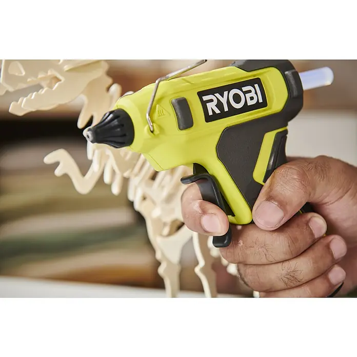 Ryobi 4V USB Akku-Heißklebepistole RGLU4-0 - 4 V, Ohne Akku, Durchmesser Klebepatrone: 11 mm - Eine Person hält eine gelb-schwarze Ryobi Heißklebepistole, bereit zum Kleben eines hellen Holz-Dinosaurierpuzzles.