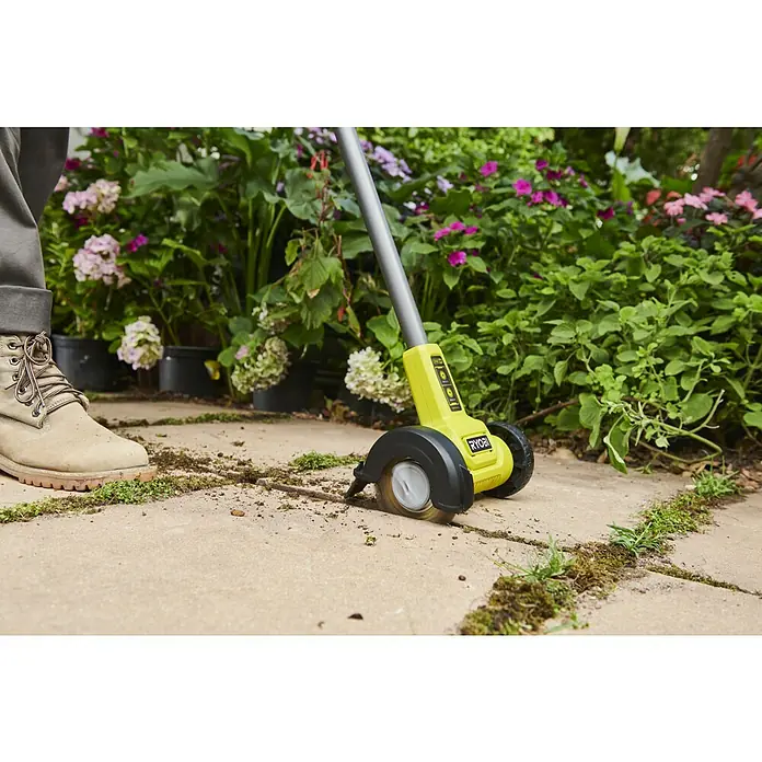 Ryobi ONE+ Akku-Fugenreiniger RY18PCC-0 - Ohne Akku - Gelb-schwarze Ryobi Unkrautbürste entfernt Moos und Unkraut aus Pflasterfugen, mit Arbeitsstiefel daneben.