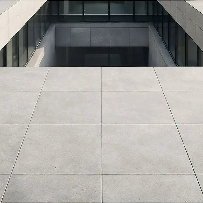 Terrassenfliese Modern Concrete - 100 x 100 x 2 cm, Grey, Matt - Hellgraue großformatige Betonfliesen bilden einen modernen Außenboden neben einem zeitgenössischen Gebäude.