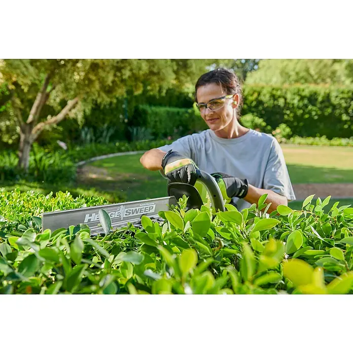 Ryobi Max Power Akku-Heckenschere RY36HT60A - 36 V, Li-Ionen, Ohne Akku, Schnittlänge: 60 cm - Person mit Schutzbrille und Handschuhen, die eine Hecke mit einer schwarz-grünen Heckenschere mit 'Hedge Sweep Debris Remover' schneidet.