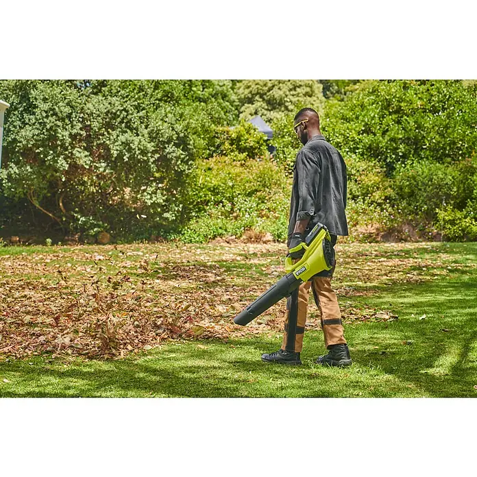 Ryobi Max Power Akku-Gebläse RY36BLB - 36 V, Ohne Akku, Blasgeschwindigkeit: 193 km/h - Eine Person im Garten bläst mit einem gelb-schwarzen Ryobi Laubbläser braune Blätter auf dem Rasen.