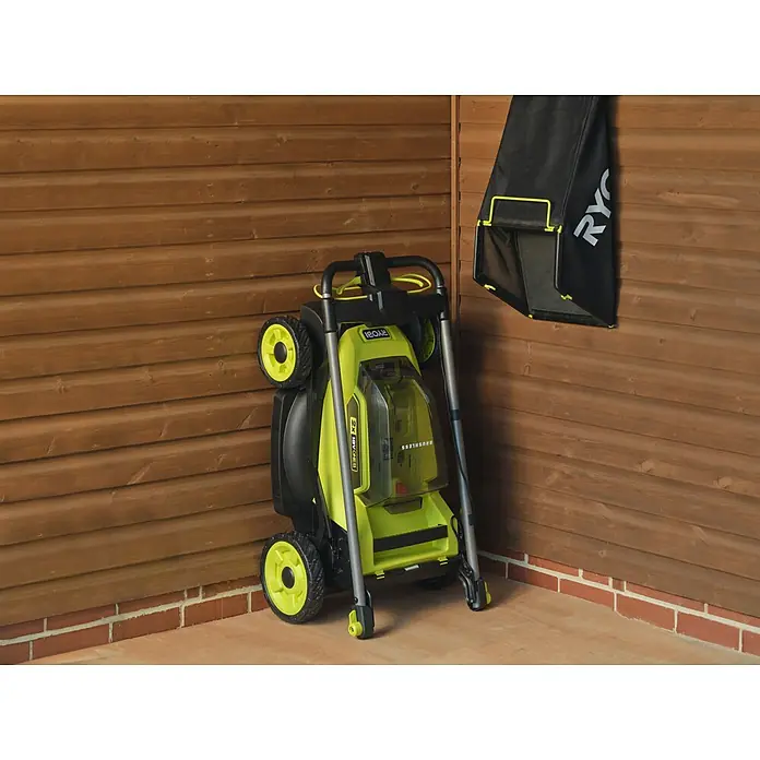 Ryobi ONE+ Akku-Rasenmäher RY18LMXSP51A - 18 V, Ohne Akku, Schnittbreite: 51 cm - Grün-schwarzer Elektrorasenmäher platzsparend zusammengeklappt, daneben hängt der schwarze Grasfangsack in einer Ecke.