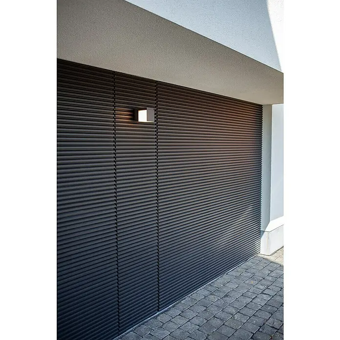 Lutec Aplique de pared LED Cuba - L x An x Al: 116 x 100 x 148 mm, Blanco neutro, Temperatura del color: 4.000 K, 500 lm - Puerta de garaje moderna de lamas oscuras con aplique de pared cuadrado iluminado encima, junto a un camino pavimentado.