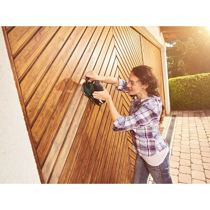 Bosch Power for All 18V Akku-Exzenterschleifer AdvancedOrbit 18 - 18 V, Li-Ionen, Ohne Akku, Durchmesser Schleifteller: 125 mm, Leerlaufdrehzahl: 3.000 U/min - 12.000 U/min - Frau schleift eine Holztür mit einem grünen Schleifgerät, Schutzbrille und kariertem Hemd für Heimwerkerprojekte im Freien.