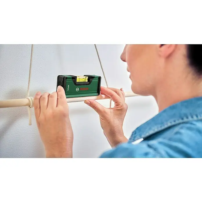 Bosch Akkuschrauber IXO 7 - 3,6 V, 1 Akku, 2 Ah, 5,5 Nm, Wasserwaagen-Set - Grünes Bosch Mini-Wasserwaage (12 cm) von Händen gehalten, um eine Holzstange auszurichten.