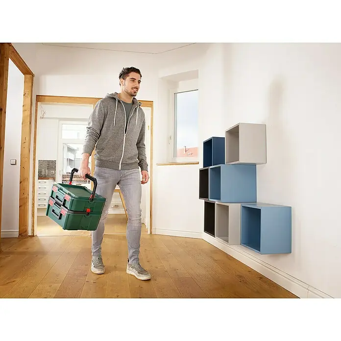 Bosch Systemkoffer M - L x B x H: 343 x 391 x 171 mm - Ein Mann trägt eine grüne Kunststoff-Werkzeugkiste mit schwarzen Griffen und roten Verschlüssen.