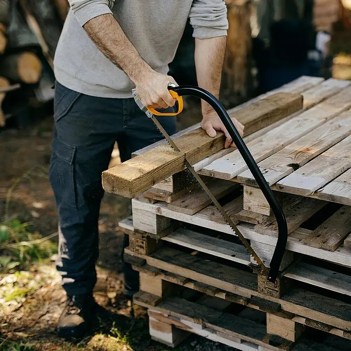 Fiskars Bügelsäge SW30 - L x H: 625 x 19 mm - Eine Person sägt mit einer Bogensäge mit schwarzem Rahmen und orangefarbenem Griff ein Holzbrett auf Paletten.