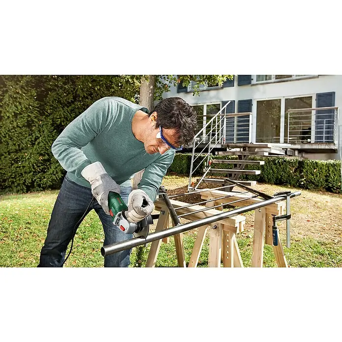 Bosch Universal Elektro-Winkelschleifer UNIVERSALGRIND 750-115 - 750 W, Durchmesser Scheibe: 115 mm, 12.000 U/min - Grüner Bosch Winkelschleifer schneidet Metallrohr; Benutzer trägt Schutzbrille und Handschuhe für Außenarbeiten.