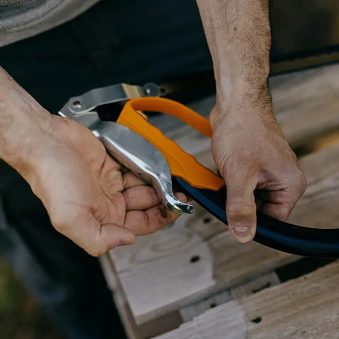 Fiskars Bügelsäge SW30 - L x H: 625 x 19 mm - Hände bedienen einen Schlauchschneider mit silbernen Klingen und orangefarbenem Griff, der einen schwarzen Schlauch schneidet.
