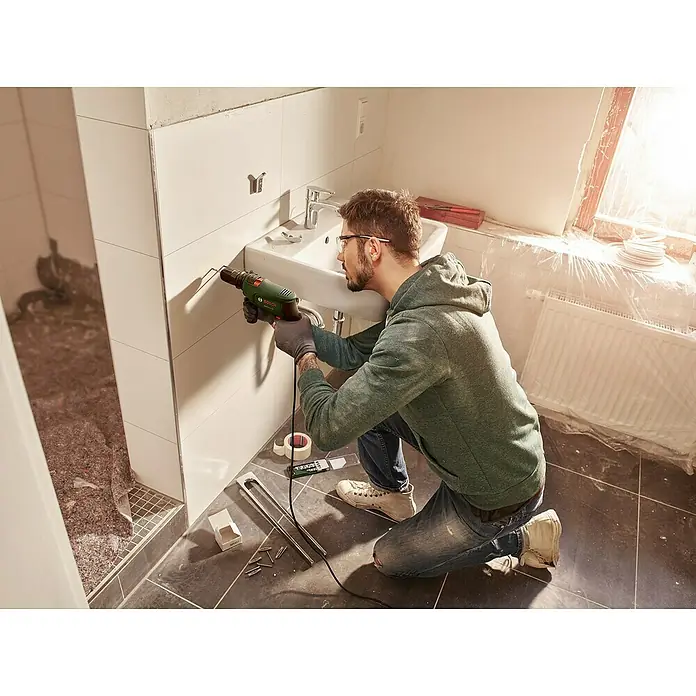 Bosch Schlagbohrmaschine EASYIMPACT 630 - 630 W, Schnellspannbohrfutter - Mann bohrt mit einem grün-schwarzen Bosch Bohrer in eine weiß geflieste Wand neben einem Waschbecken.