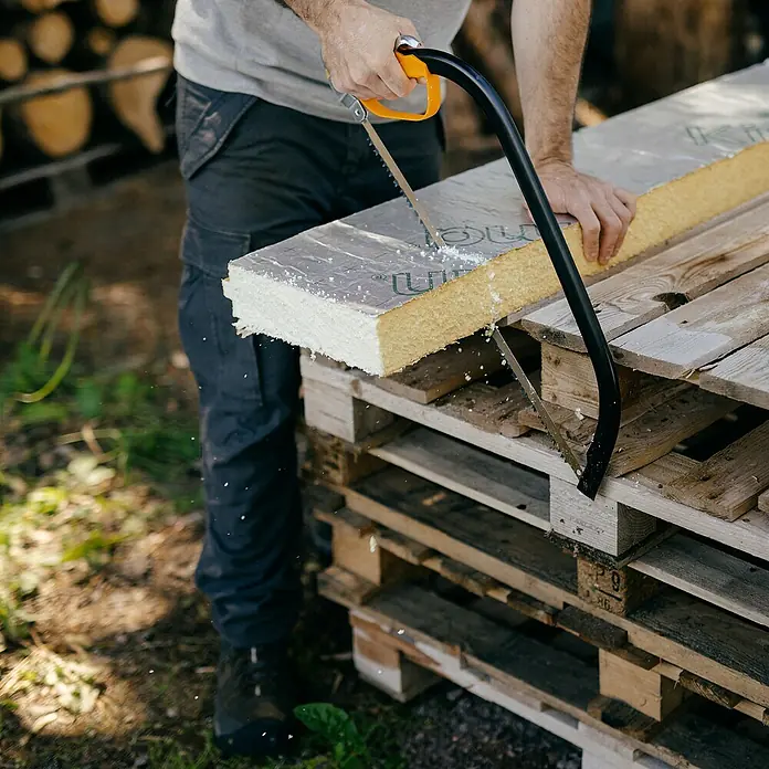 Fiskars Bügelsäge SW30 - L x H: 625 x 19 mm - Person schneidet eine gelb-weiße, in Folie verpackte Dämmplatte mit einer Handsäge.