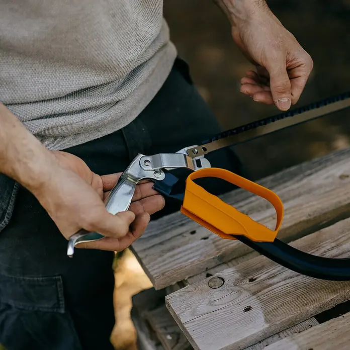 Fiskars Bügelsäge SW30 - L x H: 625 x 19 mm - Nahaufnahme von Händen, die eine silberne Klappsäge mit orangefarbenem Griff über Holzbrettern halten.