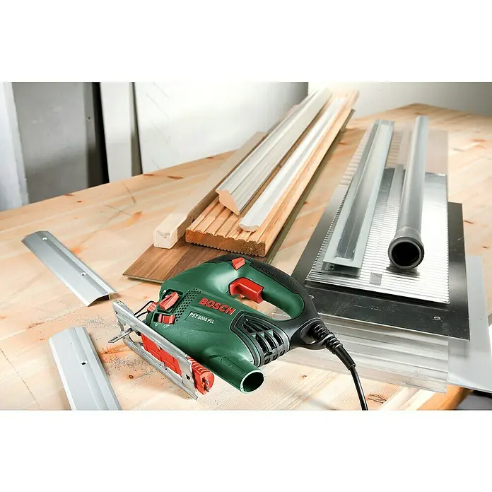 Bosch Elektro-Stichsäge PST 8000 PEL + 5TLG. SAEGEBLATT - 290 W - 530 W, 500 /min - 3.000 /min, 80 mm - Grüne Bosch PST 8000 PEL Stichsäge auf Werkbank, umgeben von Holzbrettern, Metallprofilen und Rohr, mit Sägespänen.
