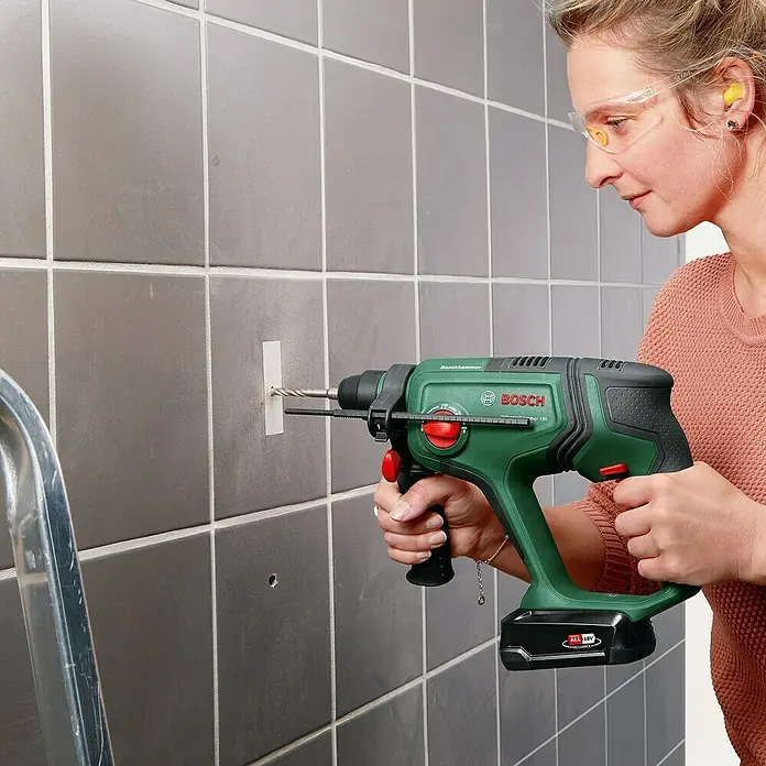 Bosch Power for All 18V Akku-Kombihammer UniversalHammer 18V - 18 V, Ohne Akku, 2 J, Mit Schnellspannbohrfutter - Frau bohrt mit einem grün-schwarzen Bosch Bohrhammer in eine geflieste Wand und trägt Schutzausrüstung.