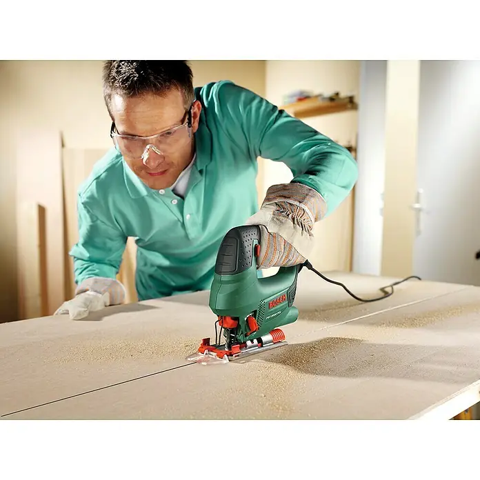 Bosch Elektro-Stichsäge PST 8000 PEL + 5TLG. SAEGEBLATT - 290 W - 530 W, 500 /min - 3.000 /min, 80 mm - Mann mit Schutzbrille und Handschuhen benutzt eine grün-schwarze Bosch Stichsäge zum Holzschneiden.