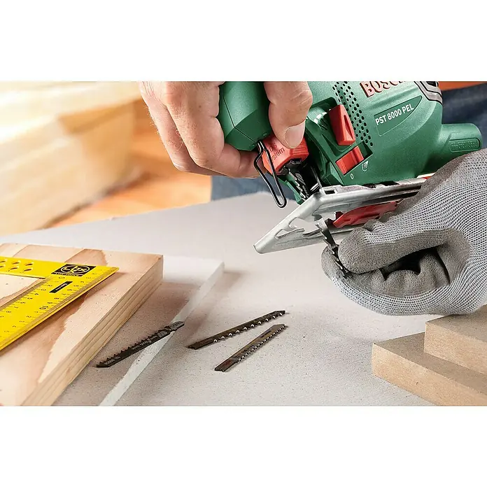 Bosch Elektro-Stichsäge PST 8000 PEL + 5TLG. SAEGEBLATT - 290 W - 530 W, 500 /min - 3.000 /min, 80 mm - Eine Person mit Handschuh wechselt ein Sägeblatt an einer grünen Bosch PST 8000 PEL Stichsäge. Verschiedene Sägeblätter und ein gelbes Lineal liegen bereit.