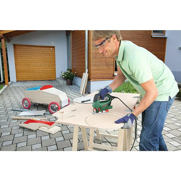 Bosch Elektro-Stichsäge PST 8000 PEL + 5TLG. SAEGEBLATT - 290 W - 530 W, 500 /min - 3.000 /min, 80 mm - Ein Mann schneidet mit einer grünen und schwarzen Bosch Stichsäge eine runde Form aus einer Holzplatte auf einer Werkbank für ein Go-Kart.