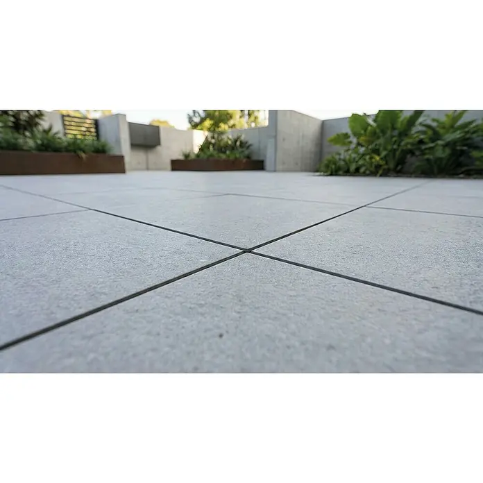 Terrassenfliese Fortezza - 60 x 60 x 2 cm, Diorit, Matt - Helle graue, strukturierte Terrassenplatten mit dunklen Fugen bilden einen Außenboden, der in die Ferne mit unscharfen Pflanzen und Betonwänden verläuft.