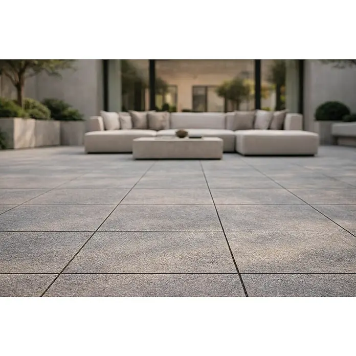 Granulati Zandobbio Terrassenfliese Fortezza - 60 x 60 x 2 cm, Diorit, Matt - Große, hellgraue, strukturierte Terrassenplatten für eine moderne Außenfläche, mit unscharfen Gartenmöbeln im Hintergrund.