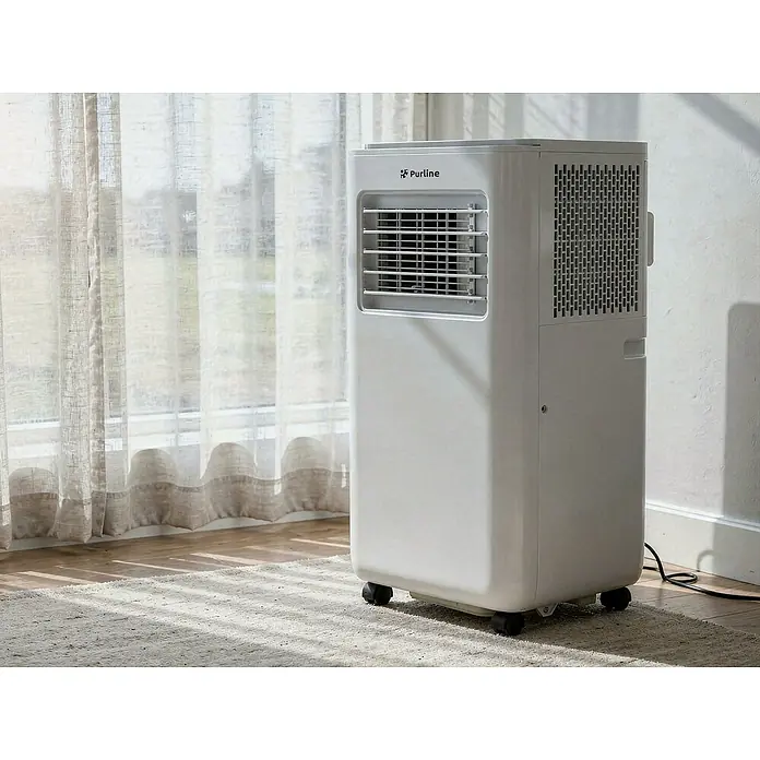 Purline Aire acondicionado portátil Cooly - 2.640 W, Tamaño de la estancia: 20 m², Blanco - Aire acondicionado portátil Purline blanco con ruedas y rejillas, colocado junto a una ventana.