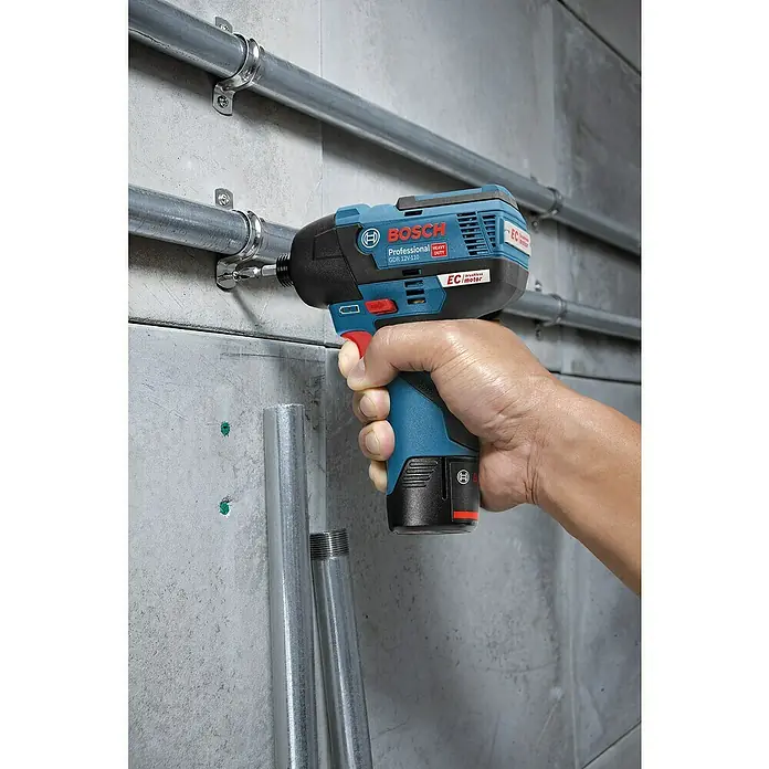 Bosch Professional AMPShare 18V Akku-Drehschlagschrauber PRO GDR 12V-110 - 12 V, 2 Akkus - Hand hält blauen und schwarzen Bosch Professional Schlagschrauber, der eine Schraube in eine Wand neben Metallrohren treibt.
