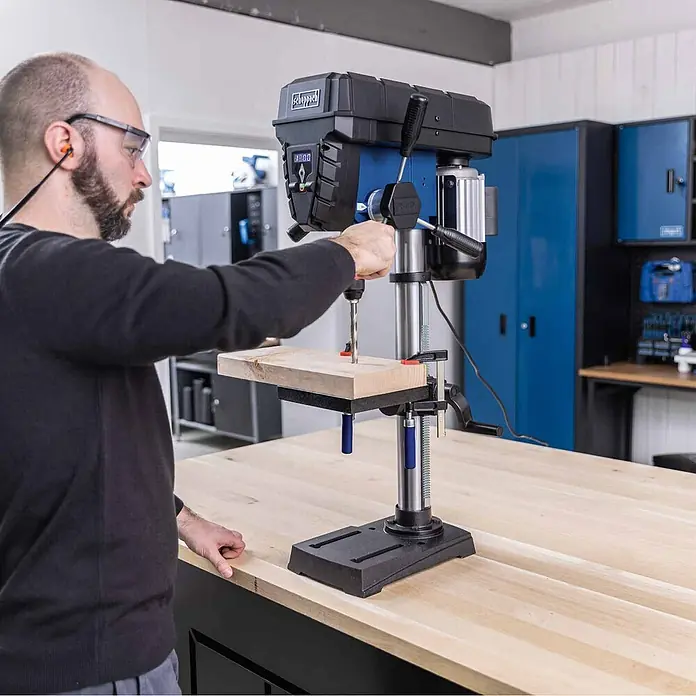 Scheppach Tischbohrmaschine TBM250 Vario - 600 W, Leerlaufdrehzahl: 1.490 U/min, Spannweite Bohrfutter: 3 mm - 13 mm - Mann bohrt mit einer blau-schwarzen Tischbohrmaschine in Holz in einer Werkstatt. Er trägt Schutzbrille und Gehörschutz.