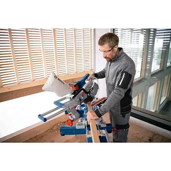 Bosch Professional AMPShare 18V Akku-Kapp- & Gehrungssäge GCM 18V-254 D - 18 V, Ohne Akku, 4.500 U/min - Mann schneidet Holz mit einer blau-silbernen Bosch Kappsäge mit Staubfangsack und Schutzbrille.