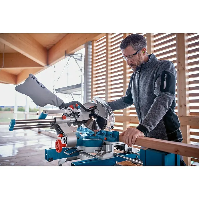 Bosch Professional AMPShare 18V Akku-Kapp- & Gehrungssäge GCM 18V-254 D - 18 V, Ohne Akku, 4.500 U/min - Mann schneidet Holzbalken mit blauer und silberner Bosch Gehrungssäge, trägt Schutzbrille und Gehörschutz.