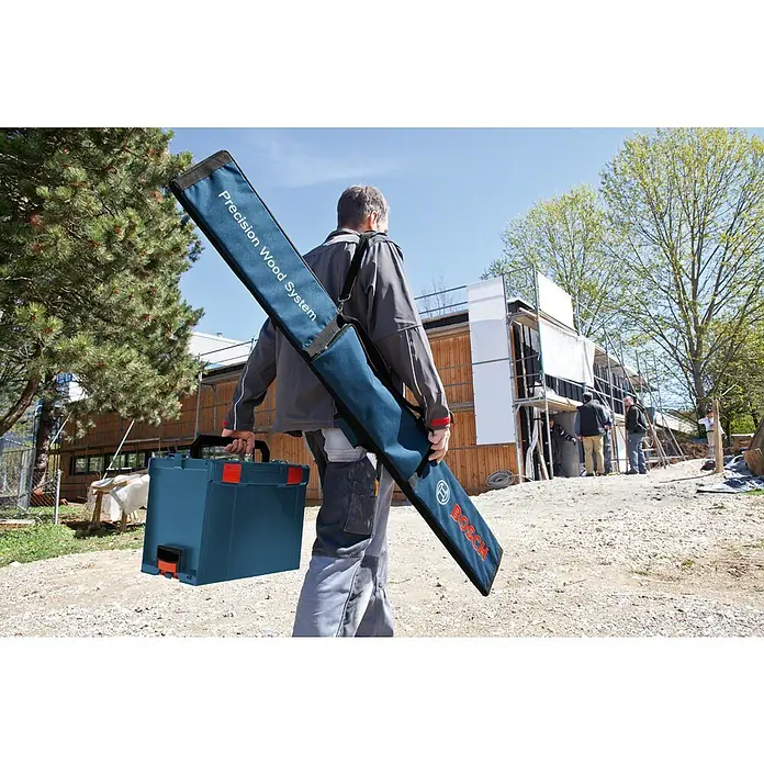 Bosch Professional Transporttasche FSN - 550 x 224 x 70 mm - Eine Person trägt eine blaue Bosch L-Boxx und eine lange blaue Stofftasche für ein Bosch 'Precision Wood System' auf einer Baustelle.