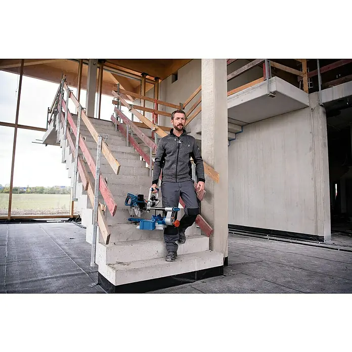 Bosch Professional AMPShare 18V Akku-Kapp- & Gehrungssäge GCM 18V-254 D - 18 V, Ohne Akku, 4.500 U/min - Mann trägt eine blau-silberne Kappsäge eine Betontreppe auf einer Baustelle hinunter.