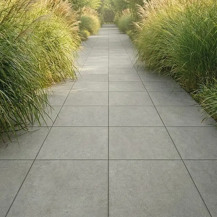 Terrassenfliese Portland - 60 x 60 x 2 cm, Londra, Matt - Graue, quadratische Pflasterplatten bilden einen Weg durch hohes Gras, der einen modernen Außenbereich schafft.