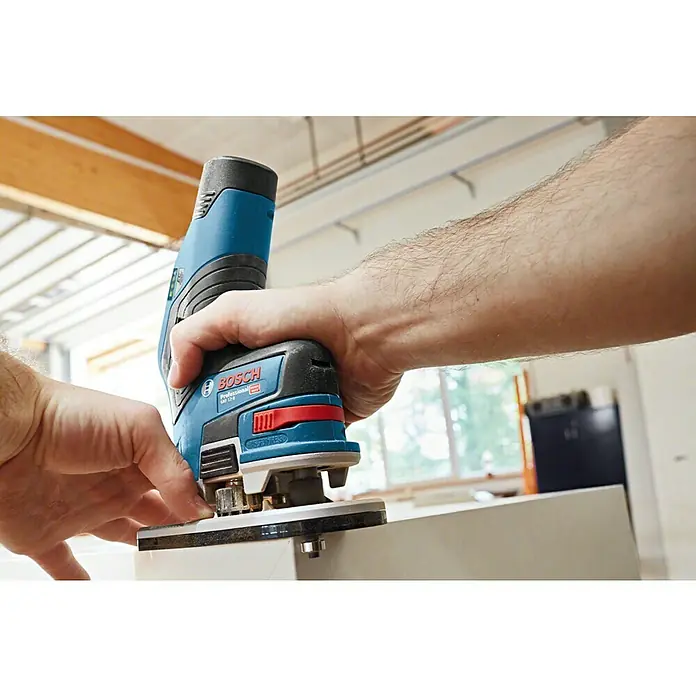 Bosch Professional Akku-Kantenfräser GKF 12V-8 PRO - 2 Akkus, 12 V - Hände bedienen eine blau-schwarze Bosch Professional Kantenfräse an einer weißen Werkstückkante.