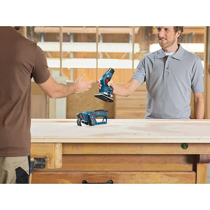 Bosch Professional Akku-Kantenfräser GKF 12V-8 PRO - 2 Akkus, 12 V - Zwei Männer in Werkstatt mit blauen Bosch Akku-Werkzeugen. Einer reicht einen Schleifer, ein Hobel liegt auf der Werkbank.