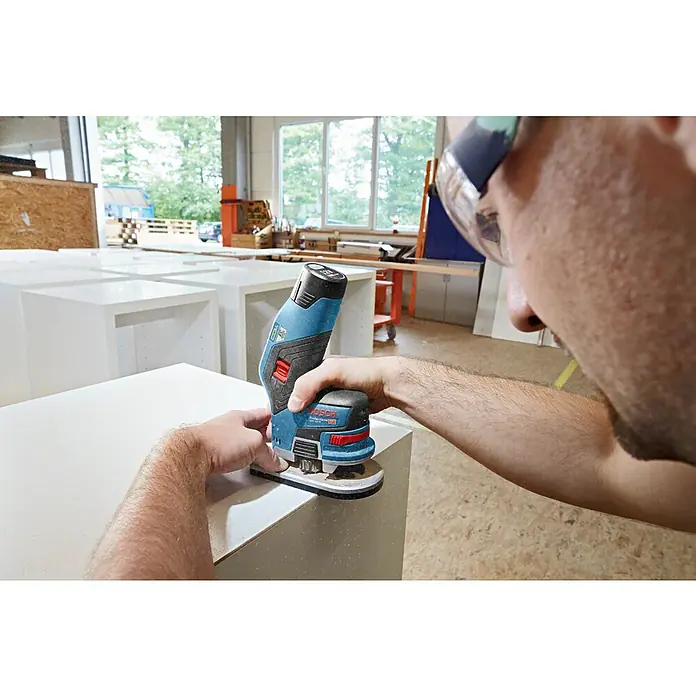 Bosch Professional Akku-Kantenfräser GKF 12V-8 PRO - 2 Akkus, 12 V - Eine Person nutzt einen blauen und schwarzen Bosch Professional Akku-Multischleifer, um die Kante eines weißen Möbelstücks zu glätten.