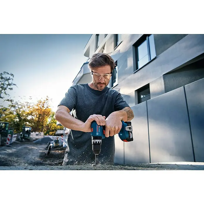 Bosch Professional AMPShare 18V Akku-Schlagbohrschrauber GSB 18V-45 PR - 18 V, 2 Akkus, 45 Nm, Inkl. L-Case Pick & Click, 82 teiliges Zub ehör-Set und 2 Akkus - Mann mit Schutzbrille bohrt mit einem blau-schwarzen Bosch Akku-Bohrschrauber in Holz auf einer Baustelle.