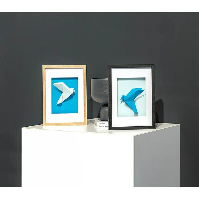 Accent Holzrahmen Objektrahmen Aura - 30 x 40 cm, Schwarz - Zwei gerahmte Origami-Vogelbilder auf einem weißen Ausstellungsblock. Eines mit hellem Holzrahmen und weißem Vogel, das andere mit dunklem Rahmen und blauem Vogel.