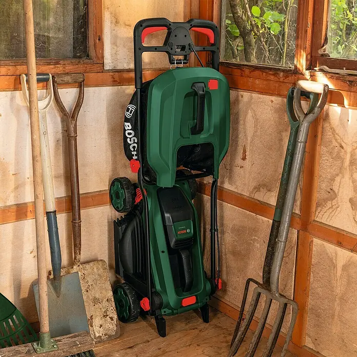 Bosch Power for All 18V Akku-Rasenmäher ROTAK18V-34 - Ohne Akku, Schnittbreite: 34 cm - Grüner und schwarzer Bosch Hochdruckreiniger aufrecht in einem Holzschuppen mit Gartengeräten.
