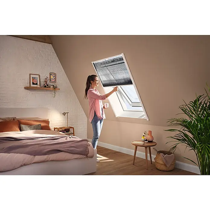 Tesa Insect Stop Insektenschutz-Dachfensterplissee teleskopierbar - Montage Insektengitter: Kleben - Frau verstellt ein dunkles Dachfensterrollo in einem hellen Dachzimmer.