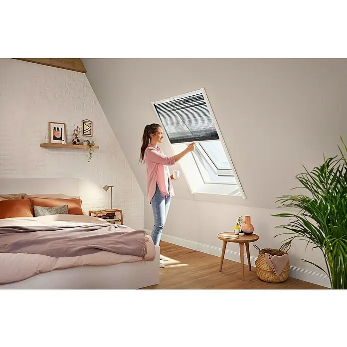 Tesa Insect Stop Insektenschutz-Dachfensterplissee teleskopierbar - Montage Insektengitter: Kleben - Eine Frau justiert ein graues Plissee an einem weißen Dachfenster in einem hellen Schlafzimmer.