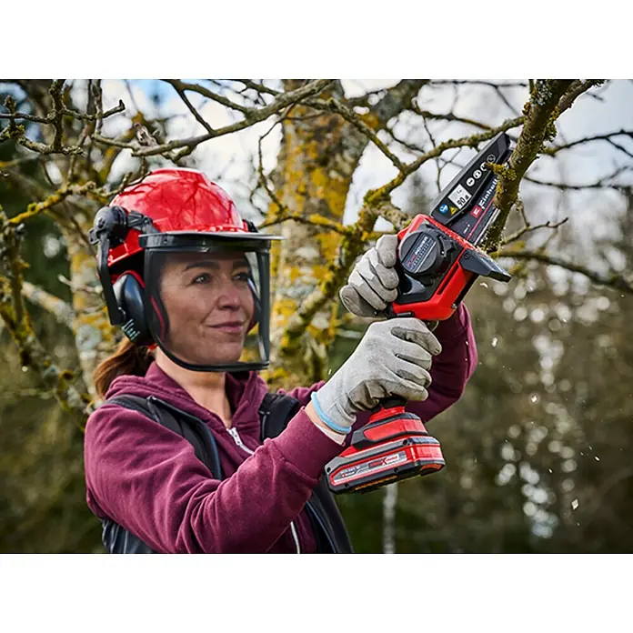 Einhell Power X-Change 18V Einhand-Akku-Kettensäge GE-PS 18/15 Li Holster Kit - 18 V, Li-Ionen, Ohne Akku, Schwertlänge: 15,8 cm - Frau mit rotem Schutzhelm schneidet Ast mit roter und schwarzer Akku-Minikettensäge; Sägespäne fliegen.