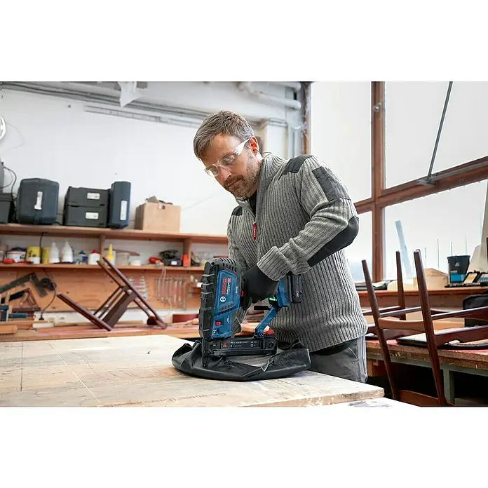 Bosch Professional Akku-Tacker GTH 18V-38 M PRO SOLO - 18 V, Ohne Akku, Klammertiefe: 10 mm - 38 mm - Mann in Werkstatt nutzt blauen und schwarzen Bosch Professional Akku-Nagler zum Befestigen von schwarzem Material, trägt Schutzbrille.