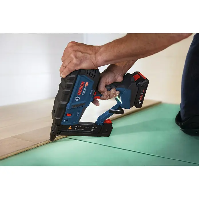Bosch Professional Akku-Tacker GTH 18V-38 M PRO SOLO - 18 V, Ohne Akku, Klammertiefe: 10 mm - 38 mm - Blau-schwarzer Bosch Akku-Nagler befestigt Holzfußboden auf grüner Unterlage.