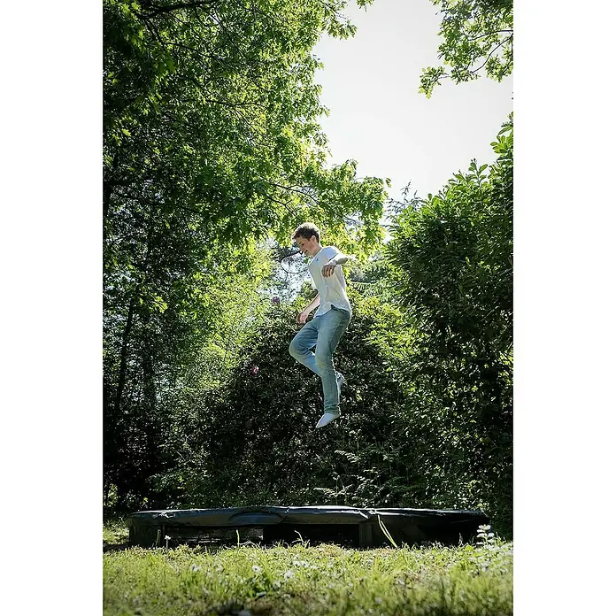 Exit Toys Trampolin Silhouette Sports - Durchmesser: 244 cm, Schwarz - Eine junge Person springt hoch auf einem runden, ebenerdigen Trampolin in einem grünen Garten. Das dunkle Trampolin fügt sich in den Rasen ein.