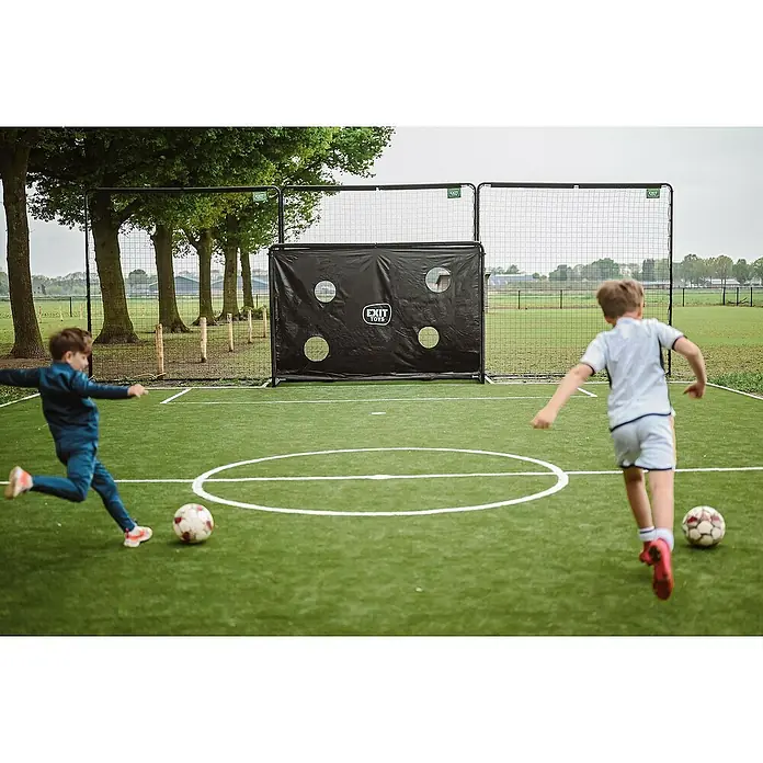 Exit Toys Sportnetz Backstop - 900 x 300 cm, Schwarz - Zwei Jungen spielen Fußball auf einem grünen Feld, einer tritt einen Ball auf ein schwarzes EXIT Toys Fußballtor mit Schießwand.
