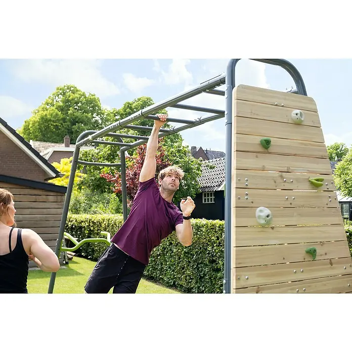 Exit Toys Klettergerüst GetSet Monkeybar MB320 - 400 x 125 x 248 cm, Stahl, Grau - Mann an grauem Metall-Klettergerüst und Holzkletterwand im Garten, Frau schaut zu. Outdoor-Fitnessgerät.