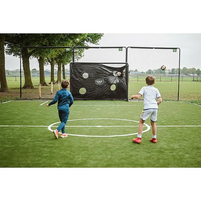 Exit Toys Sportnetz Backstop - 900 x 300 cm, Schwarz - Zwei Jungen spielen Fußball mit einem schwarzen EXIT TOYS Zielnetz auf einem Kunstrasenplatz.