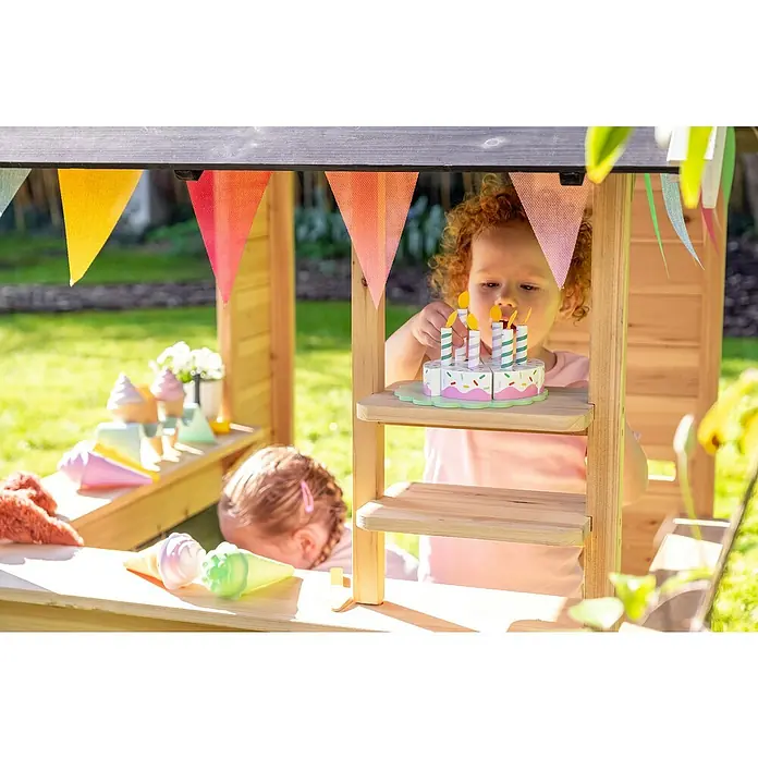 Exit Toys Spielhaus Hika - L x B x H: 140 x 111 x 155 cm, Holz, Natur - Kind spielt in einem Holzspielhaus mit bunter Girlande und schmückt einen Spielzeugkuchen mit Kerzen.